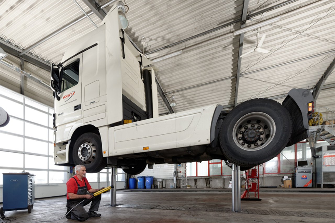 Weißer LKW auf Hebebühne, darunter ein Mechaniker in rotem T-Shirt mit gelber Sprühflasche.