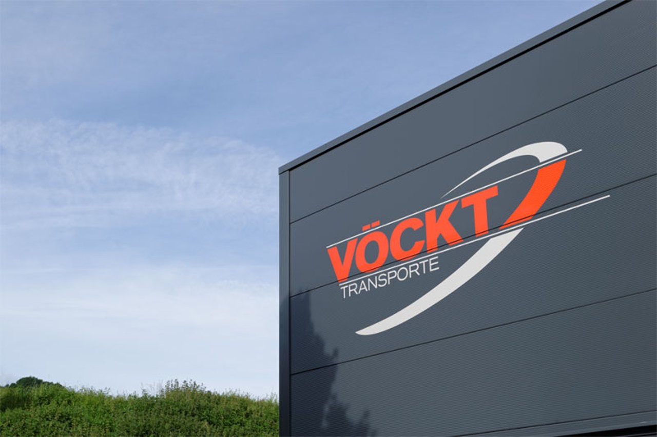 VÖCKT TRANSPORTE Firmenlogo auf einer dunkelgrauen Gebäudewand unter einem blauen Himmel.
