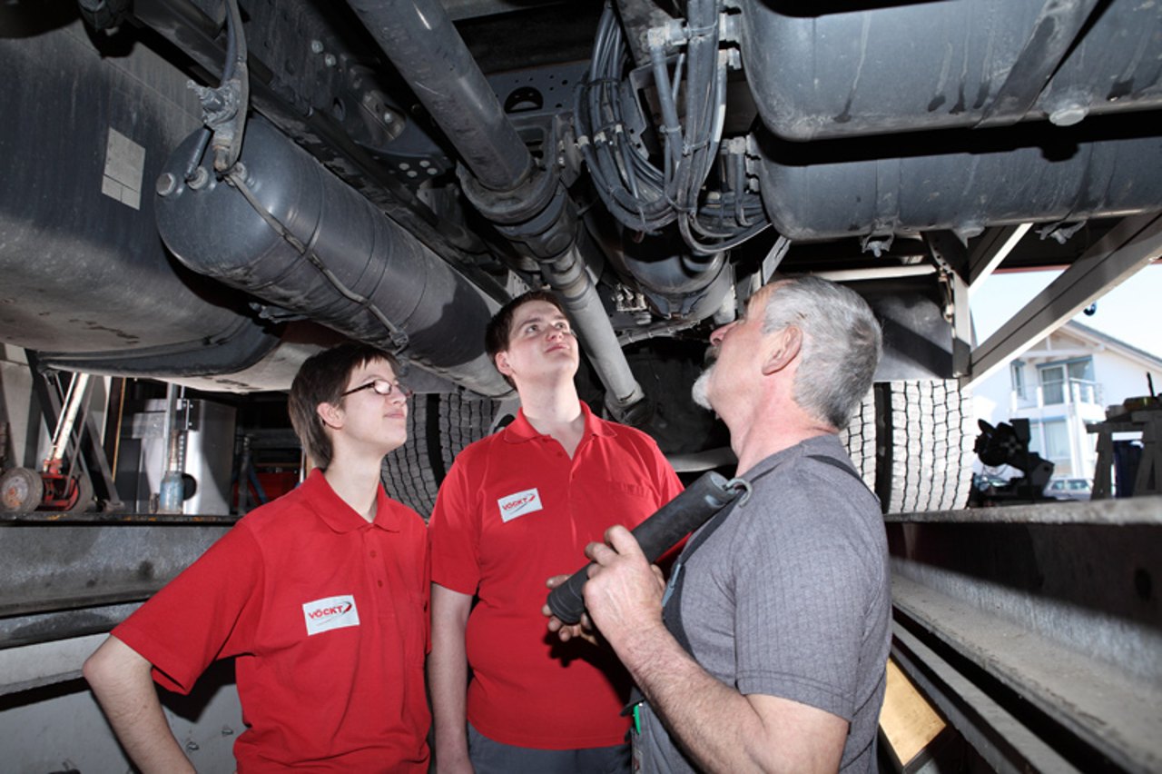 Zwei junge Männer in roten Poloshirts beobachten einen erfahrenen Mechaniker, der mit einer Taschenlampe den Unterboden eines Lastwagens untersucht.