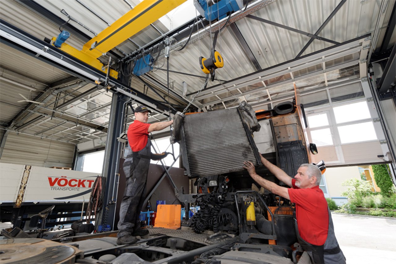 Zwei Mechaniker heben mit einem Kran einen großen Kühler in einen Lastwagen in einer Werkstatt.