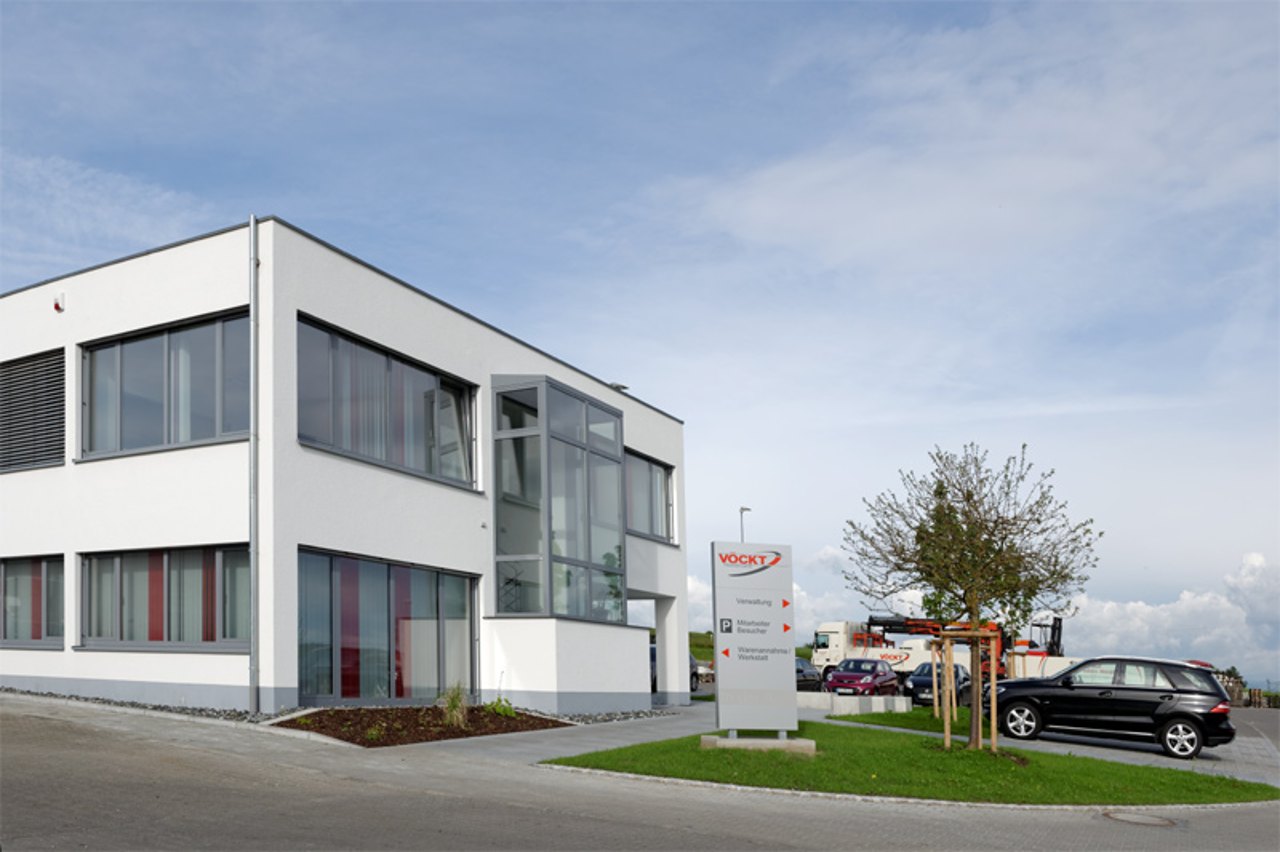 Modernes, weißes Firmengebäude mit Schild "KOCHT" und Parkplatz mit schwarzem Auto.