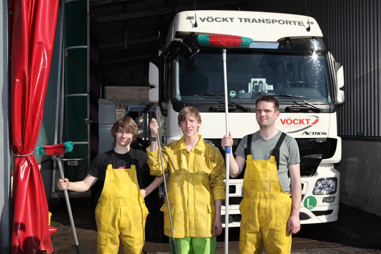 Drei junge Männer in gelben Regenhosen und Latzhosen halten Reinigungsbürsten vor einem weißen VÖCKT TRANSPORTE LKW.